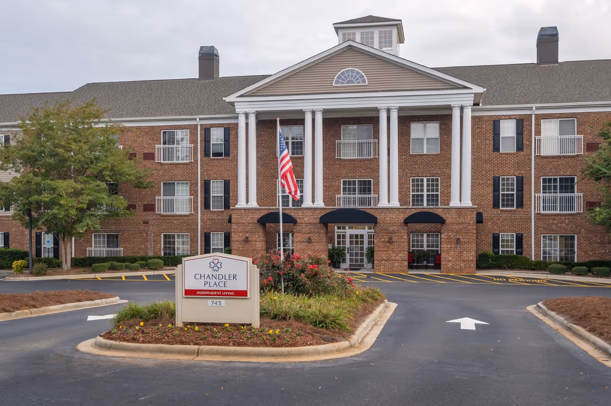 Front exterior view of Chandler Place Independent Living facility, a three-story brick building with white columns at the entrance, an American flag, and a landscaped roundabout with flowers and shrubs.