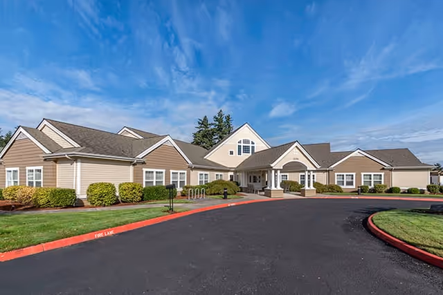 Exterior view of a single-story senior living facility building with beige siding, white trim, and a covered entrance. The building is surrounded by neatly trimmed bushes and a well-maintained lawn under a blue sky with scattered clouds. A paved driveway with red curbs leads to the entrance.