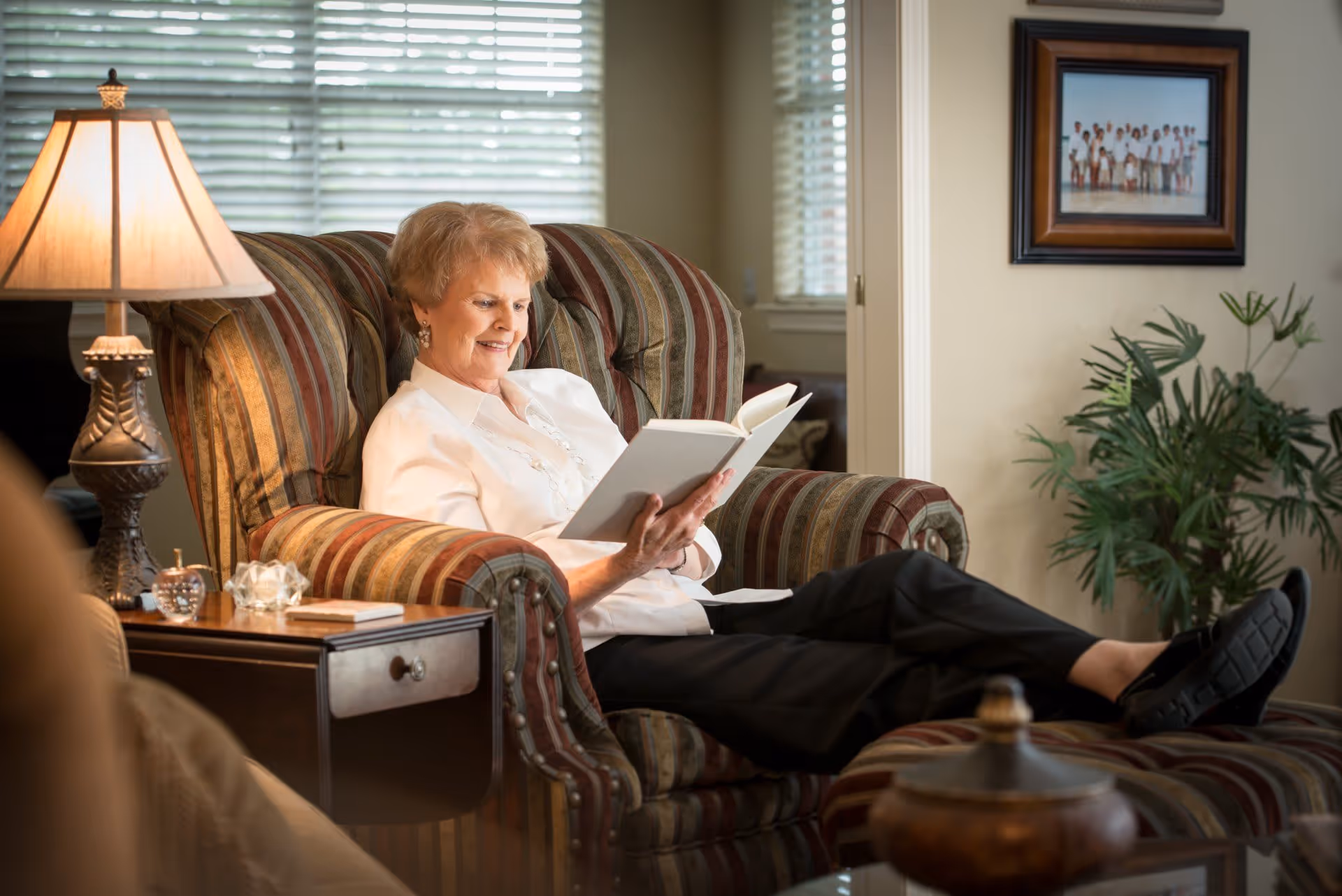 An elderly woman is sitting comfortably in a striped armchair with her feet resting on a matching ottoman. She is reading a book and smiling. The room is warmly lit by a table lamp on a side table next to her. There is a framed family photo on the wall and a green potted plant nearby.