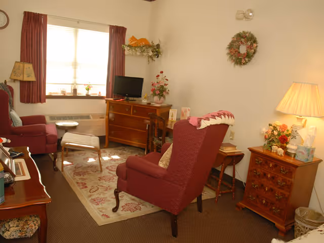 Sunlit common living room with burgundy armchairs, wooden dressers and side tables, a small TV, lamp, and floral decorations.