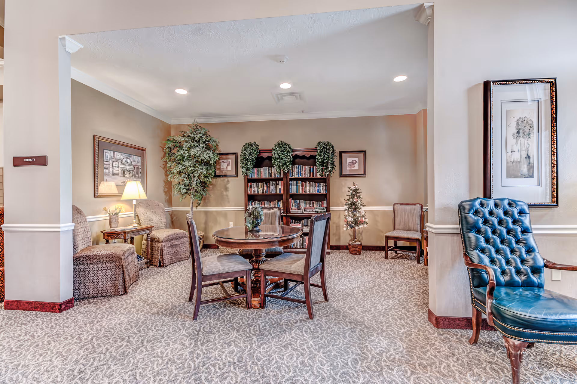 A cozy library room in a senior living facility with a round wooden table and four chairs in the center. There are two upholstered armchairs with a side table and lamp on the left, a bookshelf filled with books and decorated with plants against the back wall, and a small decorated Christmas tree. A blue tufted leather chair is positioned on the right side near a framed artwork on the wall. The room has beige walls, patterned carpet, and recessed ceiling lights.