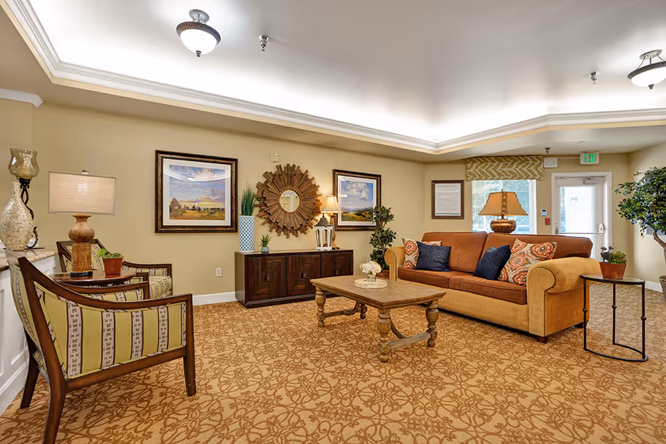 A cozy living room area with a brown and orange sofa adorned with patterned and solid pillows, a wooden coffee table with a small floral arrangement, two wooden armchairs with striped cushions, a dark wooden cabinet with decorative items including a round sunburst mirror and framed landscape paintings on the wall, and warm lighting from table lamps and ceiling fixtures. The room has beige walls and a patterned carpet, with a door and window in the background.