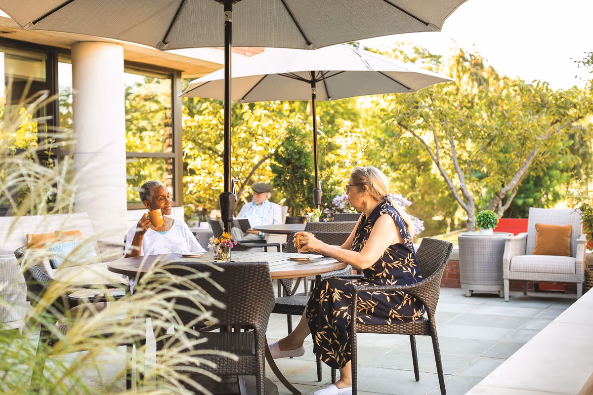 Two women sit at a round patio table under large umbrellas, enjoying drinks on a sunny outdoor terrace surrounded by plants and seating.