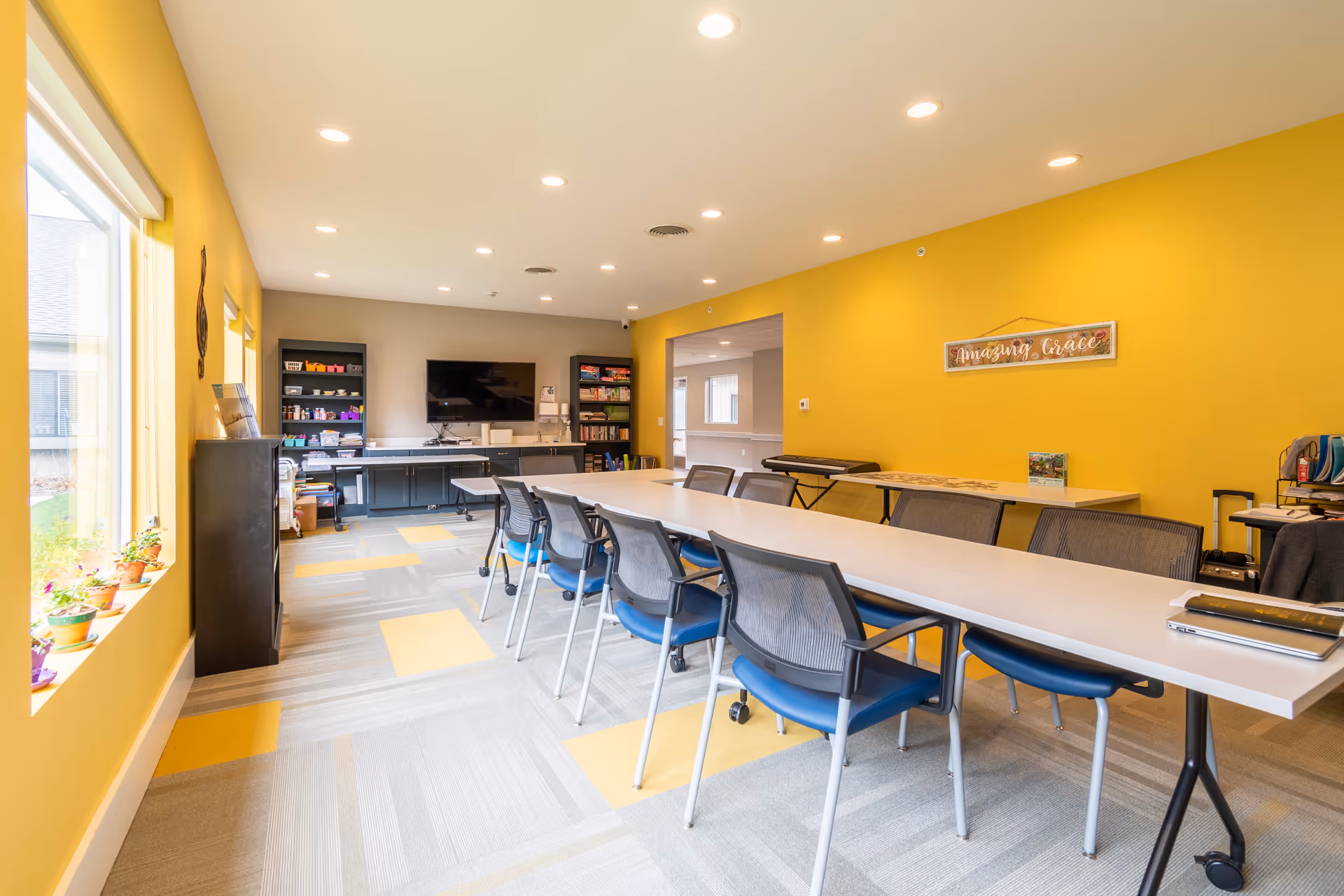A bright and spacious activity room with yellow walls and a long white table surrounded by blue and black chairs. The room has large windows with potted plants on the windowsill, a TV mounted on the far wall, shelves with games and supplies, and a sign on the wall that reads 'Amazing Grace'.