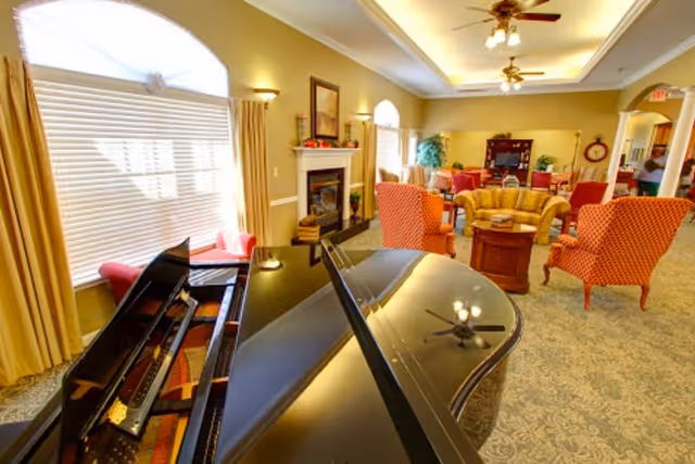 A bright and spacious senior living common area featuring a black grand piano in the foreground, comfortable seating including orange armchairs and a yellow sofa arranged around a wooden coffee table, a fireplace with a framed picture above it, large windows with yellow curtains, ceiling fans, and warm lighting.