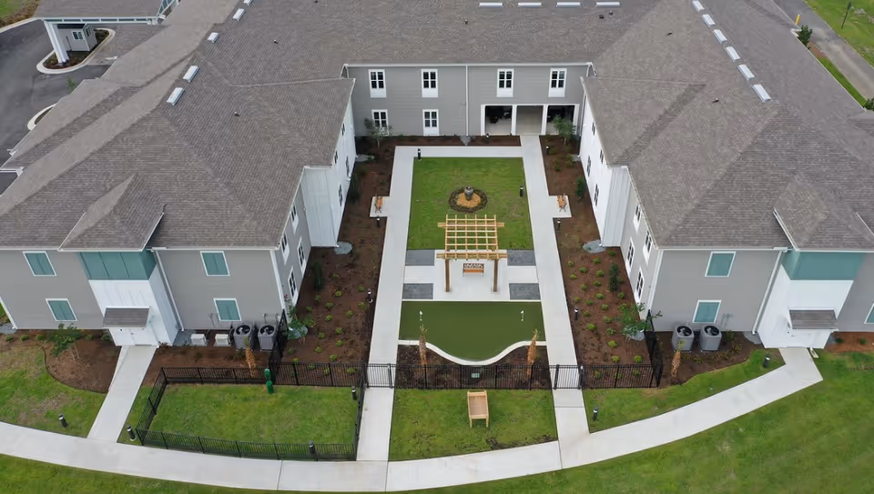 Aerial view of The Canopy at Walden Woods senior living facility showing a U-shaped building surrounding a landscaped courtyard with a small putting green, a wooden pergola, benches, and walking paths.