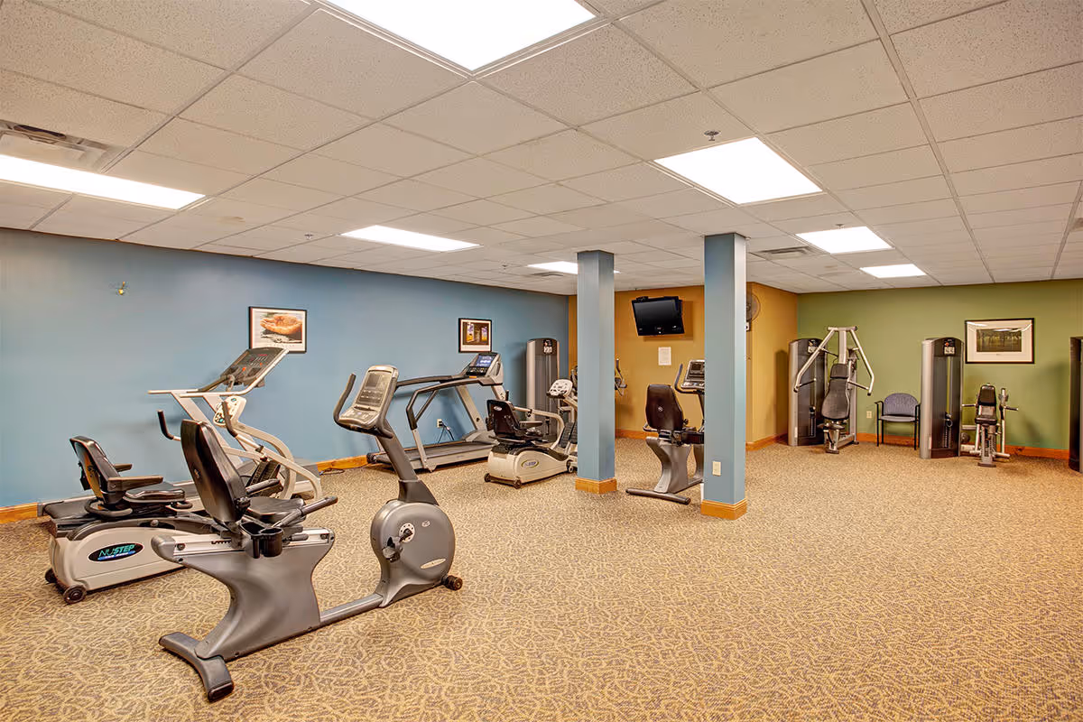 A fitness room with various exercise equipment including stationary bikes, treadmills, and weight machines. The room has a carpeted floor, blue and green painted walls, framed pictures, and a wall-mounted TV.