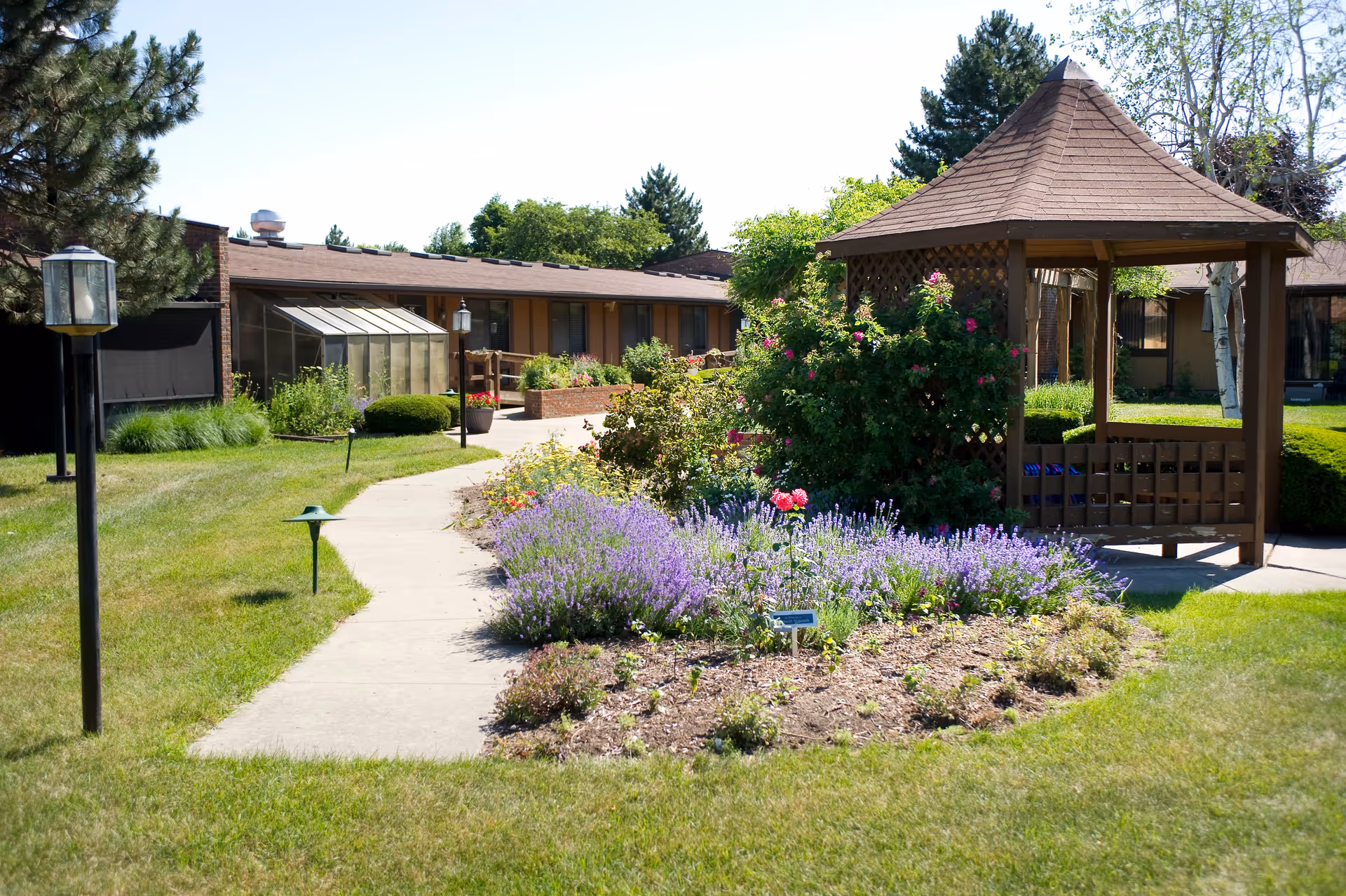 A sunny outdoor garden area at The Village of Westland featuring a paved walkway, a wooden gazebo, colorful flower beds with purple and pink flowers, green grass, and a building with large windows in the background.