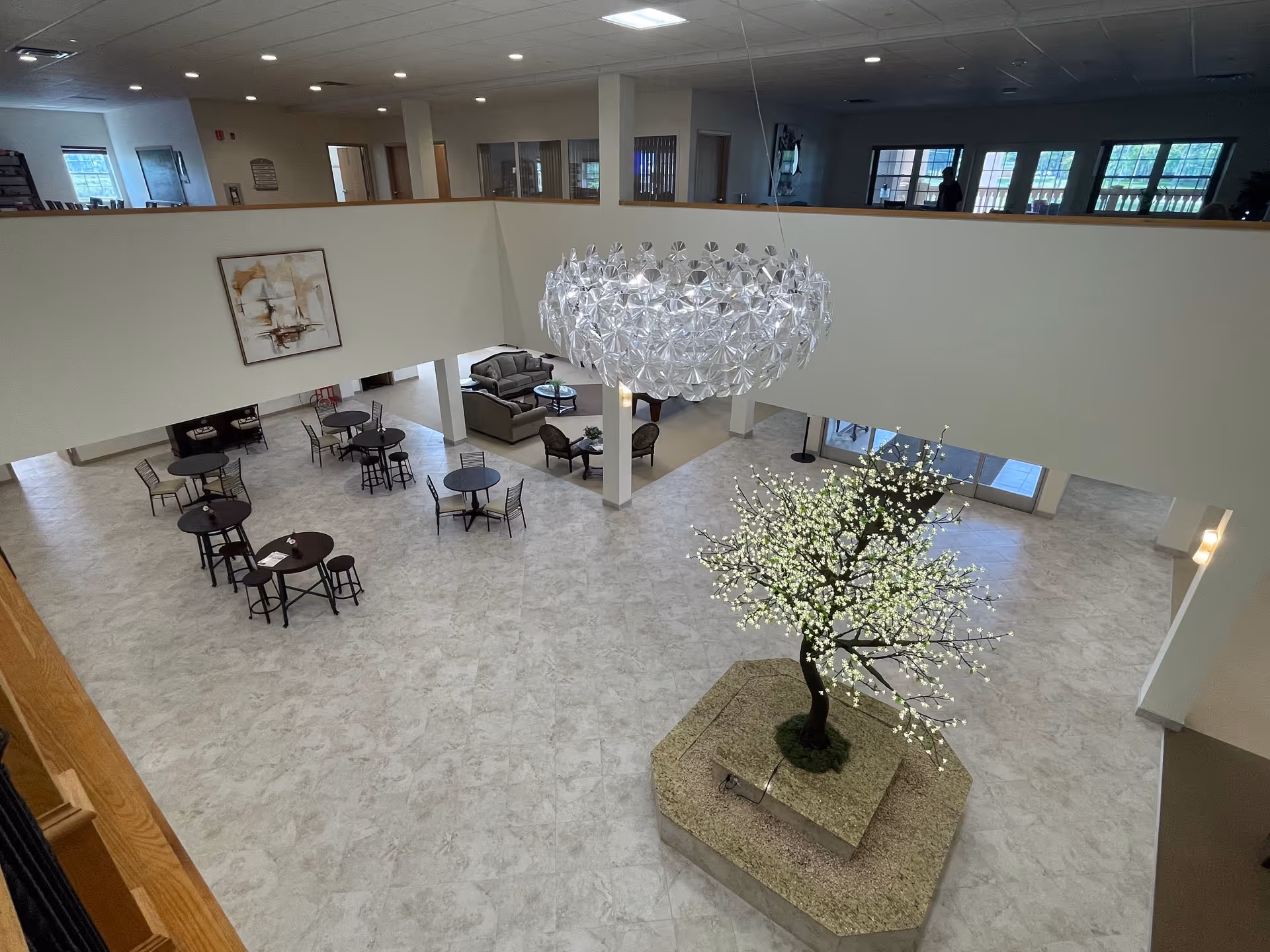 Interior view of a spacious senior living facility common area with a large decorative lighted tree centerpiece on a raised platform, several round tables with chairs, a seating area with sofas and armchairs, a modern chandelier hanging from the ceiling, and large windows letting in natural light.