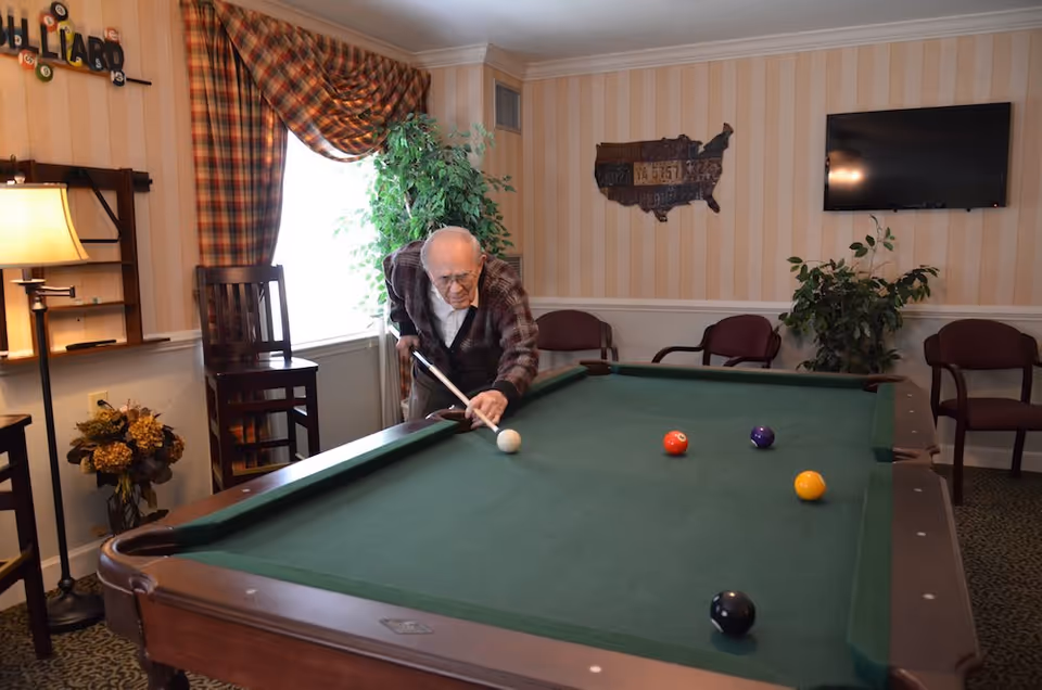 An elderly man playing pool in a cozy room with striped wallpaper, a window with plaid curtains, several chairs, a floor lamp, and a wall-mounted TV. There is a decorative wooden map of the United States on the wall and some plants in the room.