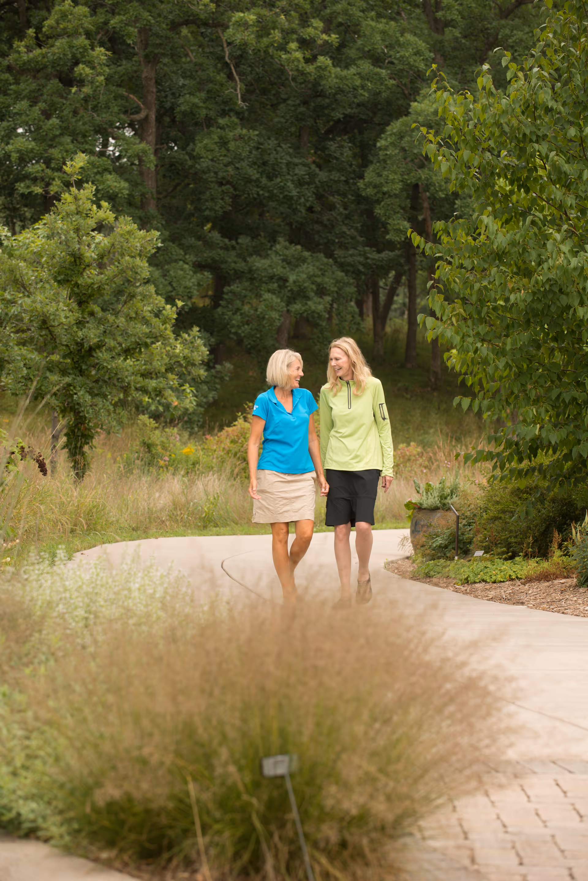 Two women walking and talking on a curved paved path surrounded by greenery and trees in an outdoor garden setting.
