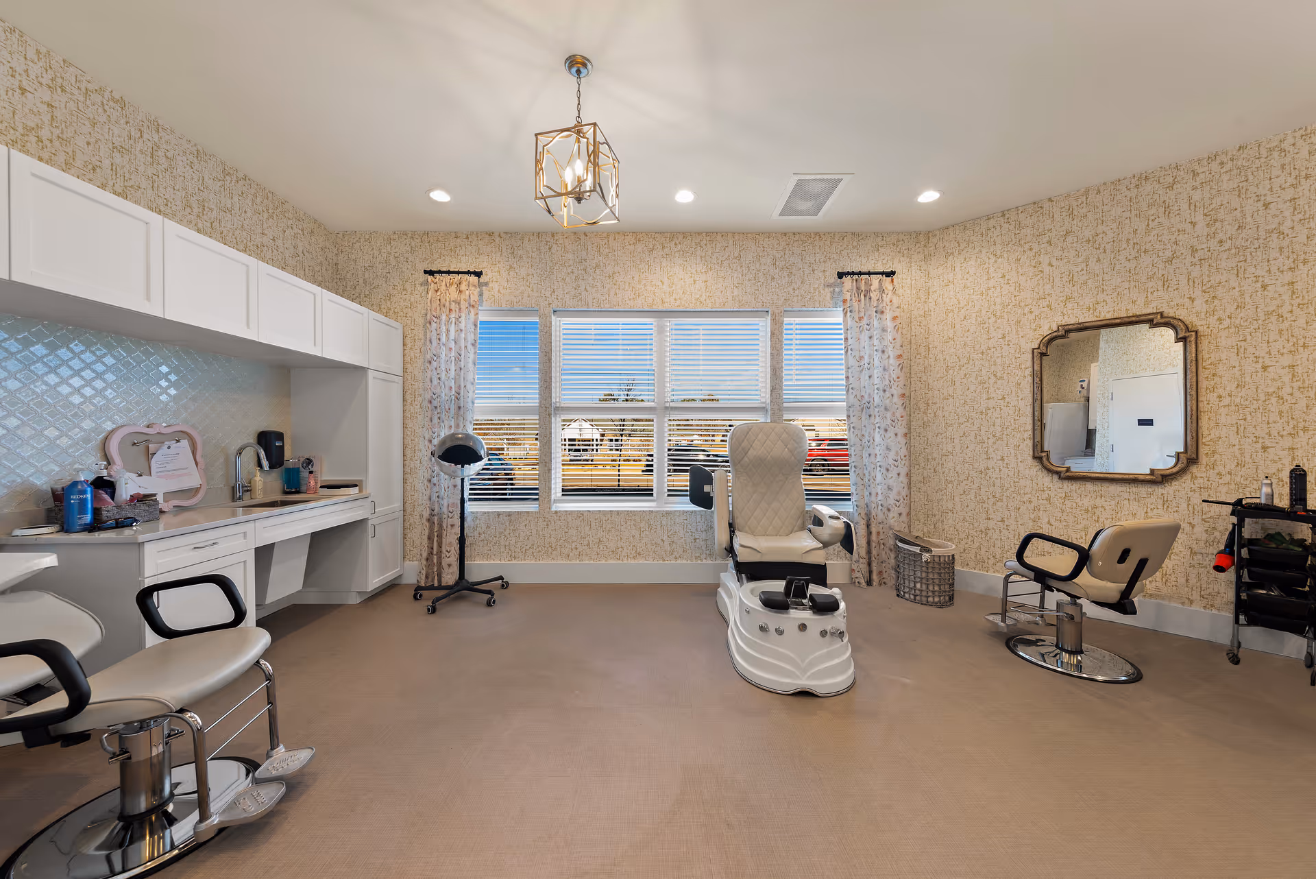 A bright and spacious salon room with beige patterned wallpaper and large windows with blinds and floral curtains. The room features a white pedicure chair in the center, two salon chairs with footrests on either side, a wall-mounted mirror, a rolling cart with salon supplies, and a white countertop with cabinets and a sink on the left side. A modern geometric light fixture hangs from the ceiling.