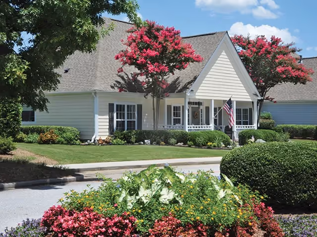 Front exterior of a single-story senior living building with a covered porch, American flag, and well-kept landscaping with flowering trees and shrubs.