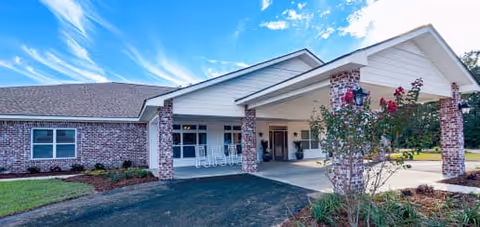 Exterior view of a single-story brick building with a covered entrance supported by brick columns. There is a driveway leading up to the entrance, landscaping with bushes and flowers, and a bright blue sky with some clouds above.