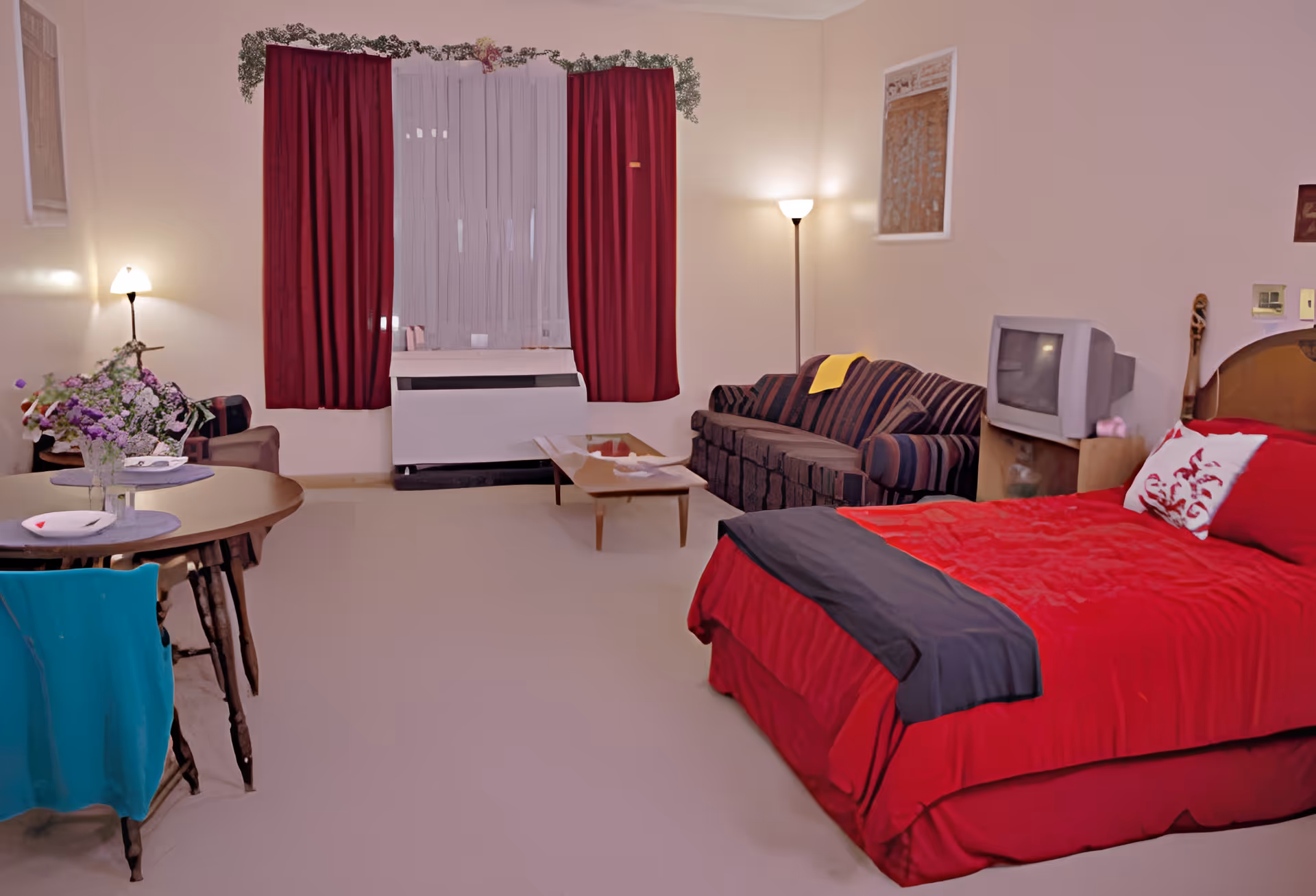 A cozy room with a bed covered in red bedding, a striped sofa, a small wooden coffee table, a round dining table with chairs, and a TV on a stand. The room has beige walls, red curtains, and two floor lamps providing warm lighting.
