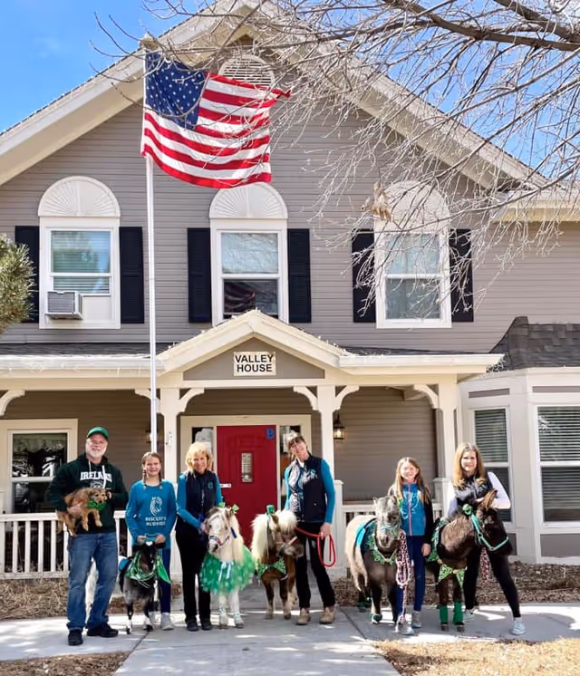 A group of six people standing in front of a two-story building with a sign that reads 'Valley House'. They are accompanied by several small ponies and a dog. An American flag is flying on a flagpole in front of the building. The people and animals appear to be posing for a photo on a sunny day.