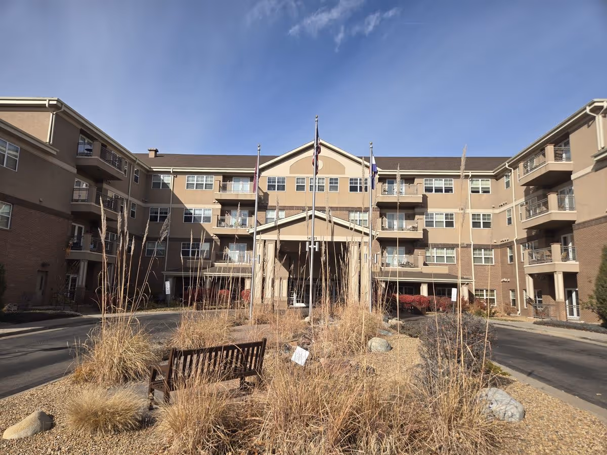 Front entrance of a multi-story senior living building with flags, balconies, landscaping and a bench in the foreground.