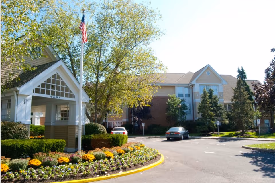 Exterior view of The Forum At Knightsbridge senior living facility showing the entrance with a covered driveway, an American flag on a flagpole, landscaped flower beds with colorful flowers, and a parking area with cars. Trees and shrubs surround the building under a clear blue sky.