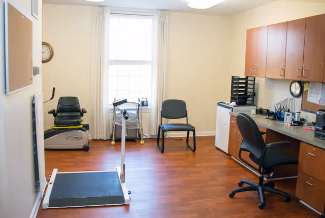 A well-lit room with wooden flooring featuring exercise equipment including a recumbent bike and a step platform. There are two chairs near a window with white curtains. On the right side, there is a desk with an office chair, cabinets above and below, and various office supplies.