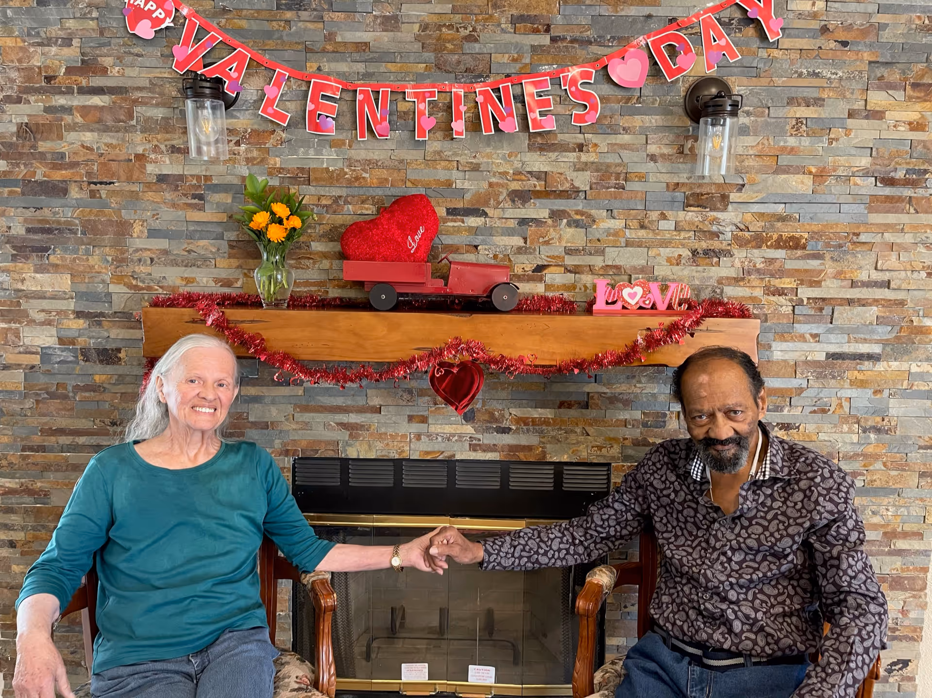 An elderly woman and man sitting on chairs in front of a stone fireplace decorated with Valentine's Day decorations, holding hands and smiling. Above the fireplace is a wooden mantle with a vase of yellow flowers, a red heart-shaped pillow, a small red toy truck, and a 'LOVE' sign. A red garland and a banner spelling 'VALENTINE'S DAY' hang on the wall.