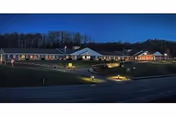 A long, single-story rehabilitation and care facility building illuminated at dusk with entrance lights, a grassy slope, and a road in the foreground.