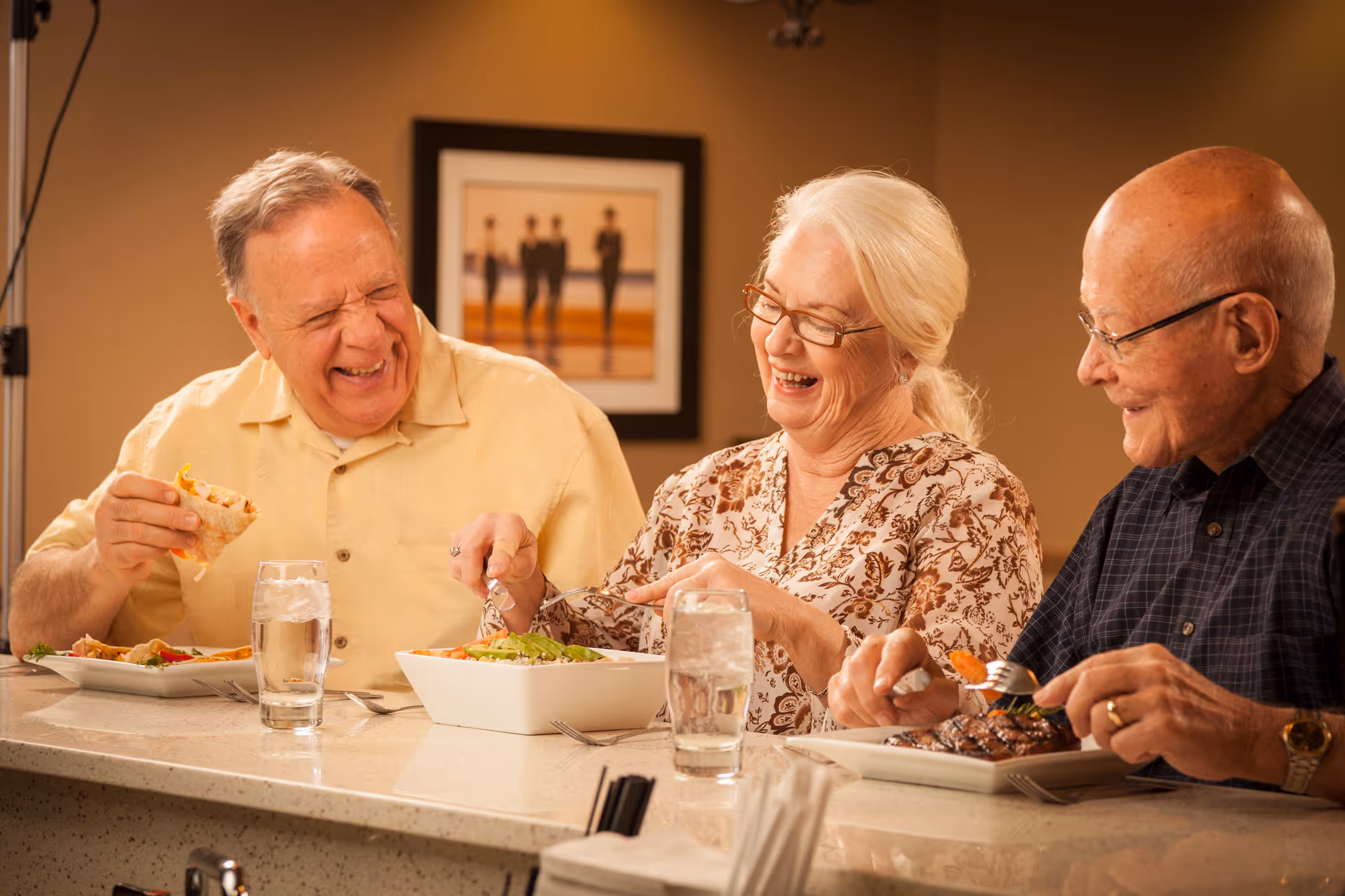 Three elderly people sitting at a counter enjoying a meal together, smiling and laughing. The man on the left is holding a wrap, the woman in the middle is serving salad from a bowl, and the man on the right is eating a meal with vegetables. There are glasses of water on the counter and a framed picture on the wall behind them.