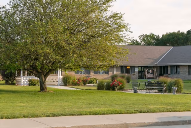 A large green lawn with a tree, benches, flowerbeds, and a single-story care facility building across the yard.