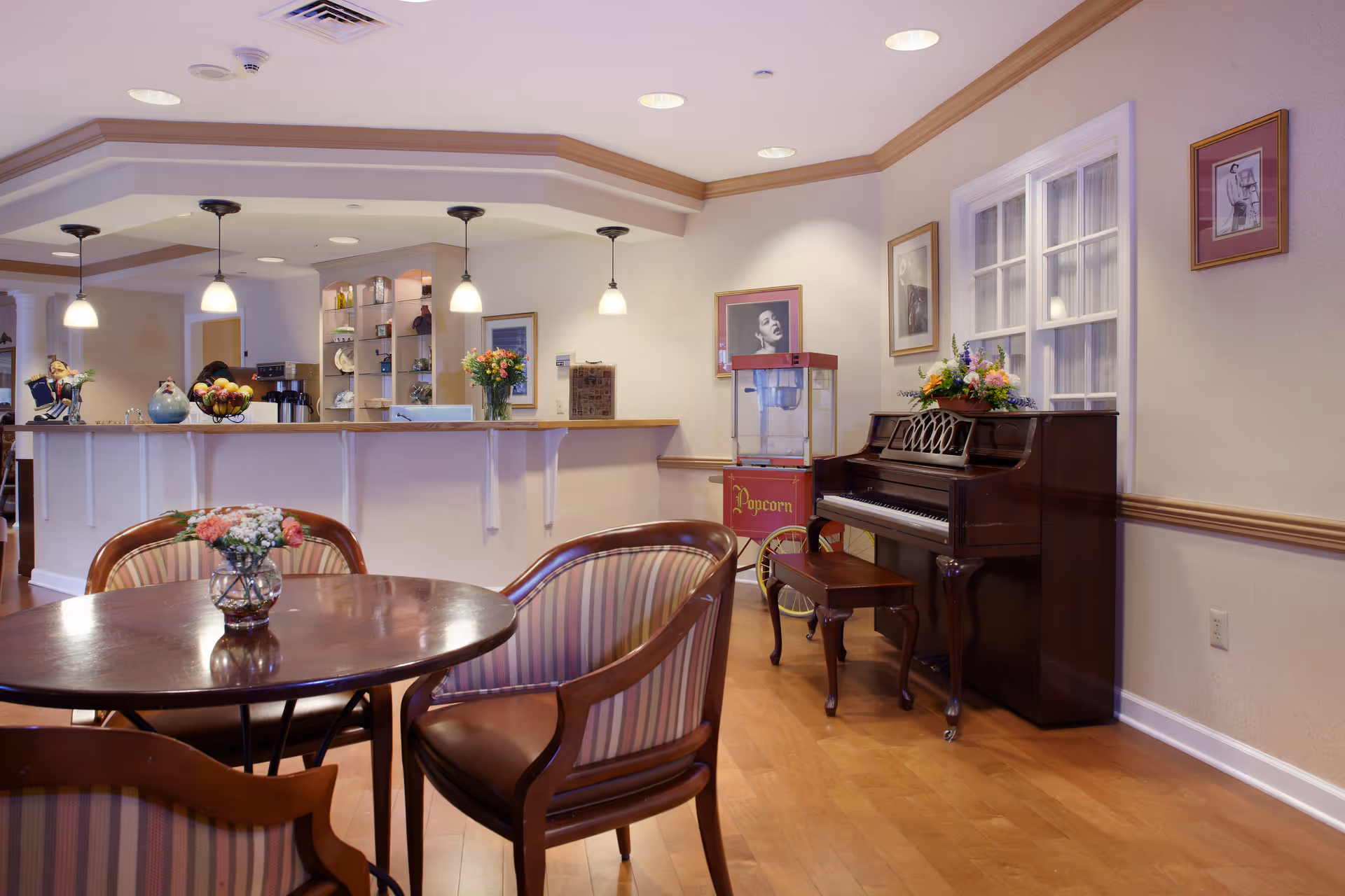 A bright senior living common area with a round table and chairs, a piano, a popcorn machine, and a service counter with pendant lights.