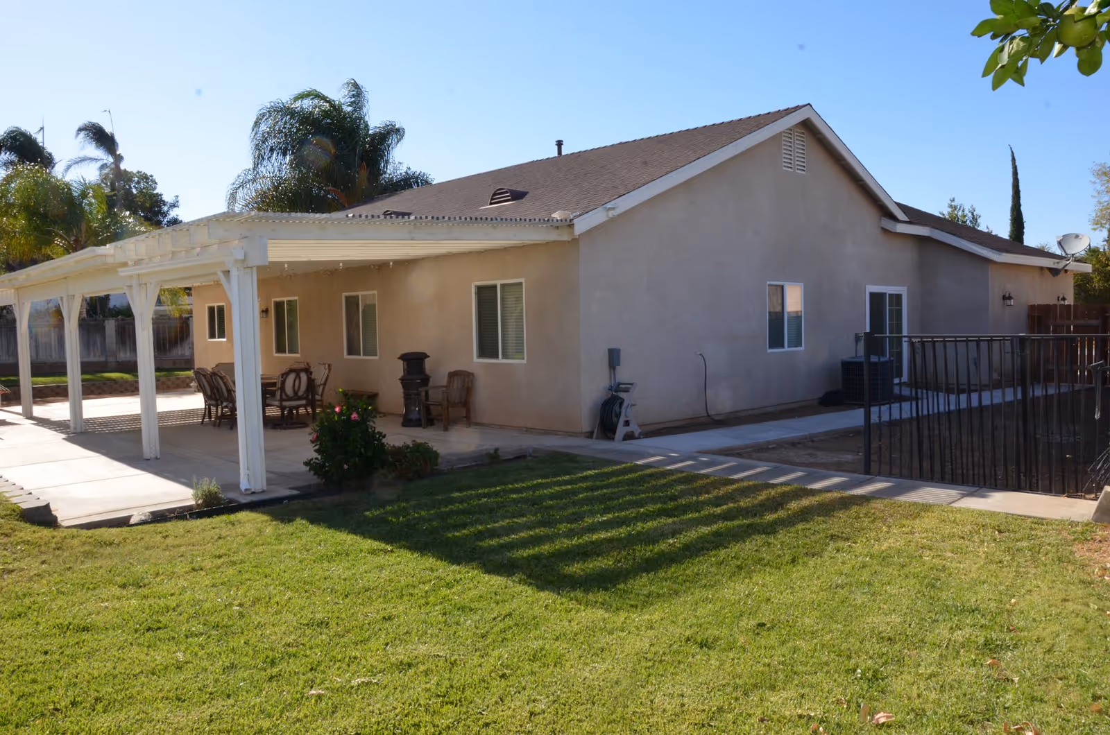 Exterior view of a single-story beige building with a brown roof, featuring a covered patio area with outdoor chairs and a table. The patio is surrounded by a well-maintained green lawn and some palm trees are visible in the background under a clear blue sky.