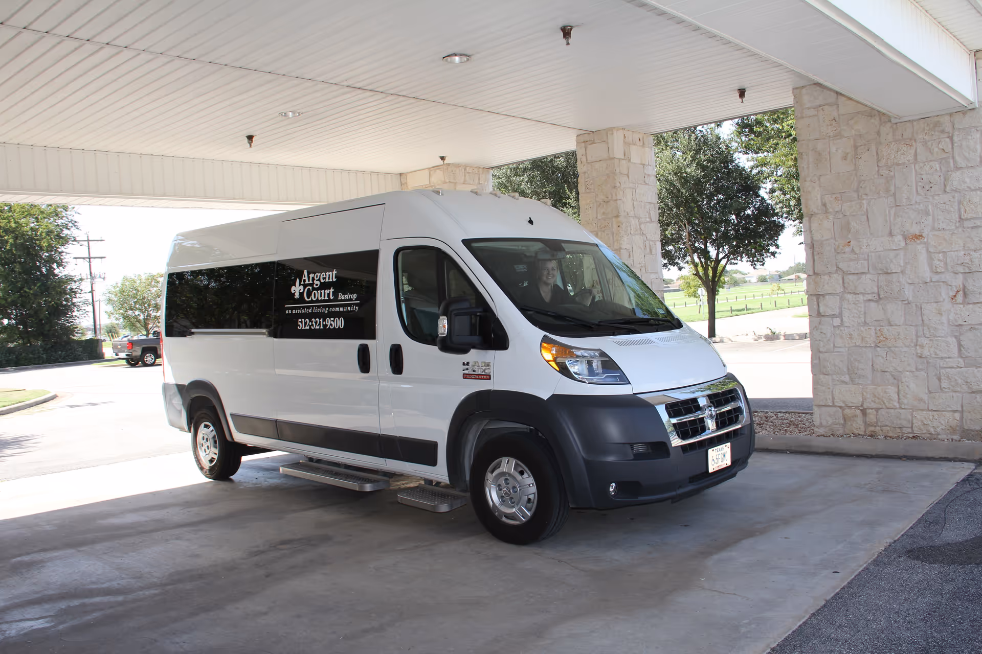 A white Argent Court Assisted Living shuttle van parked under a covered entrance beside stone pillars.