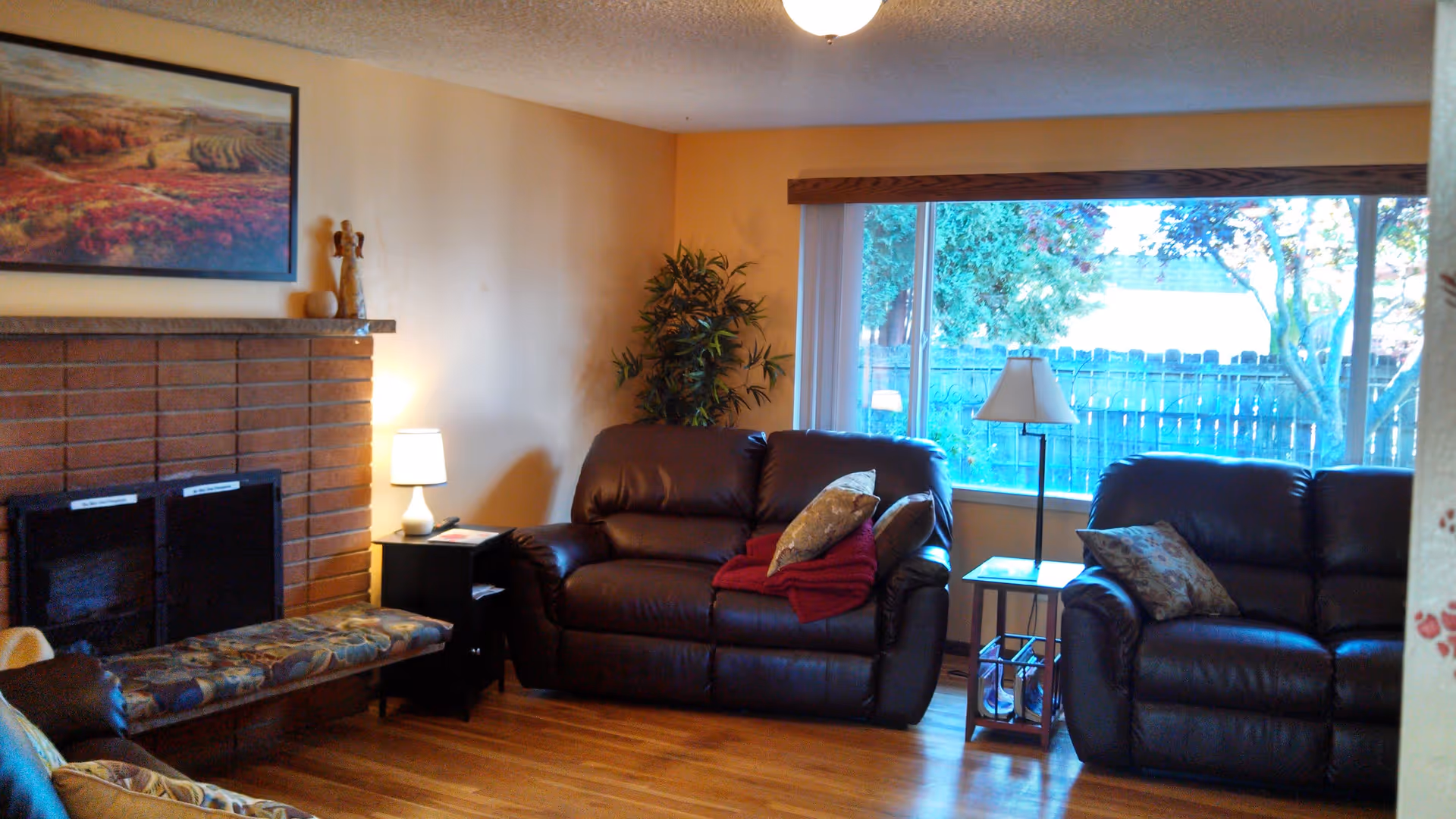 Cozy living room with two dark brown leather sofas, a wooden floor, a large window with a view of a fenced backyard with trees, a brick fireplace with a floral cushioned bench in front, a small side table with a lamp, and a framed landscape painting above the fireplace.