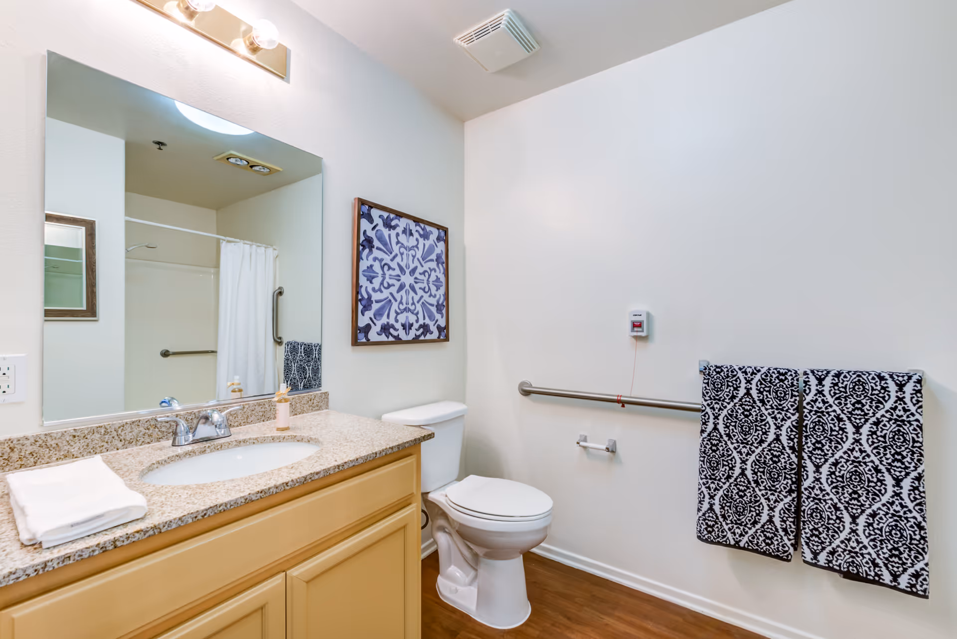 A clean and well-lit bathroom with a beige vanity featuring a granite countertop and an oval sink. Above the vanity is a large mirror with a light fixture. To the right is a white toilet with a safety grab bar on the wall behind it. Two patterned black and white towels hang on a towel rack next to the toilet. A framed decorative artwork is mounted on the wall above the toilet. The floor is wooden, and the walls are painted white.