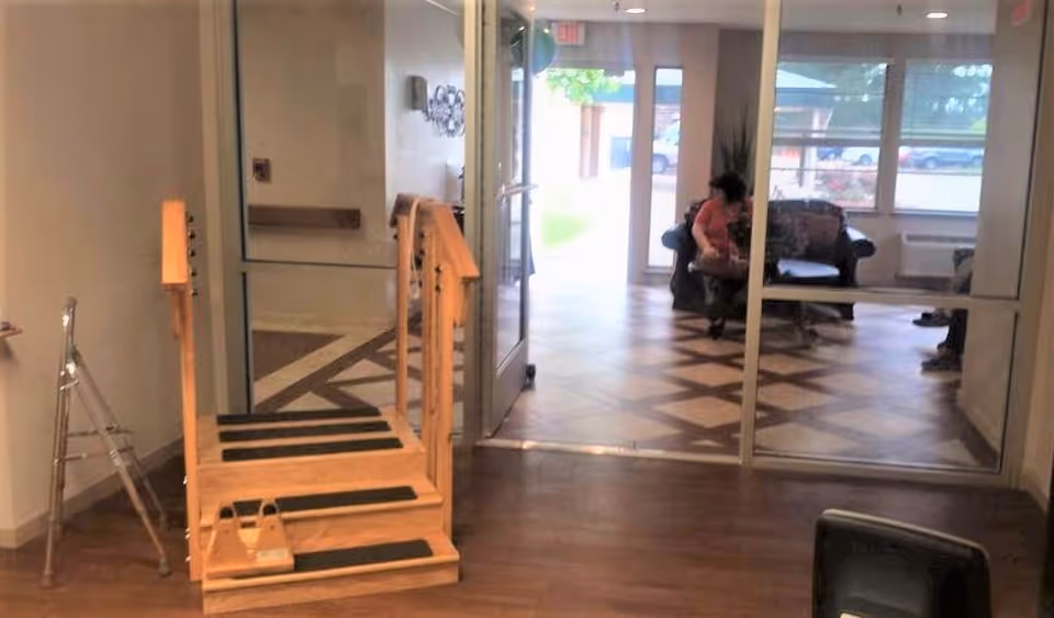 Interior view of a rehabilitation center showing a wooden physical therapy step with handrails and a walker nearby. Beyond a glass door, there is a seating area with a person sitting on a couch and large windows letting in natural light.