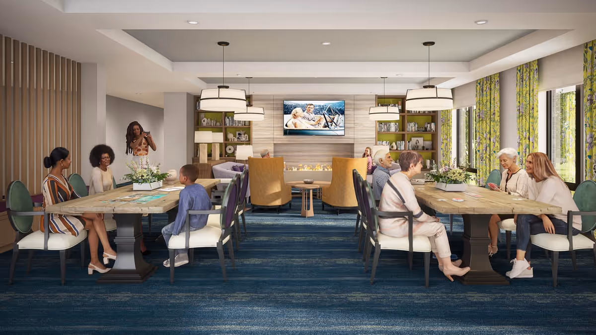 A bright communal dining and lounge area with long tables, people seated and conversing, pendant lights, a wall-mounted TV, and large windows.
