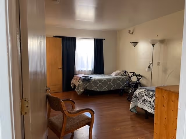 A simple bedroom with two single beds, one covered with a gray patterned blanket and the other with a white and black patterned blanket. There is a wooden chair with armrests near the door, a wooden dresser, a walker next to one of the beds, and a floor lamp with two light fixtures. The room has wooden flooring and a window with dark curtains letting in natural light.
