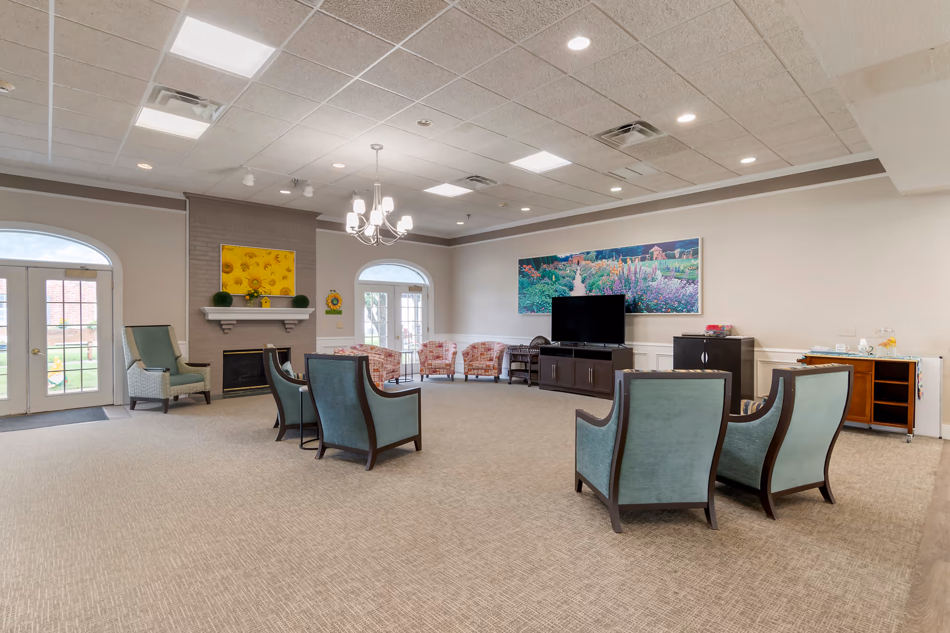 A spacious and well-lit common living area in a memory care community featuring multiple upholstered armchairs arranged in a semi-circle facing a flat-screen TV on a dark wooden cabinet. The room has a beige carpet, a chandelier, recessed lighting, and two sets of French doors with arched windows leading outside. A fireplace with a mantle decorated with small plants and a sunflower welcome sign is visible on one wall. A large colorful painting of a garden hangs above the TV. A small serving cart with refreshments is placed against the wall.