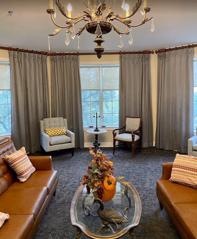 A cozy living room with two brown leather sofas, each with patterned pillows, two armchairs, and a small round table between the windows. The table holds a decorative scale. In the center is a glass coffee table with a fall-themed floral arrangement featuring a pumpkin and leaves. The room has large windows with gray curtains and a decorative chandelier hanging from the ceiling.