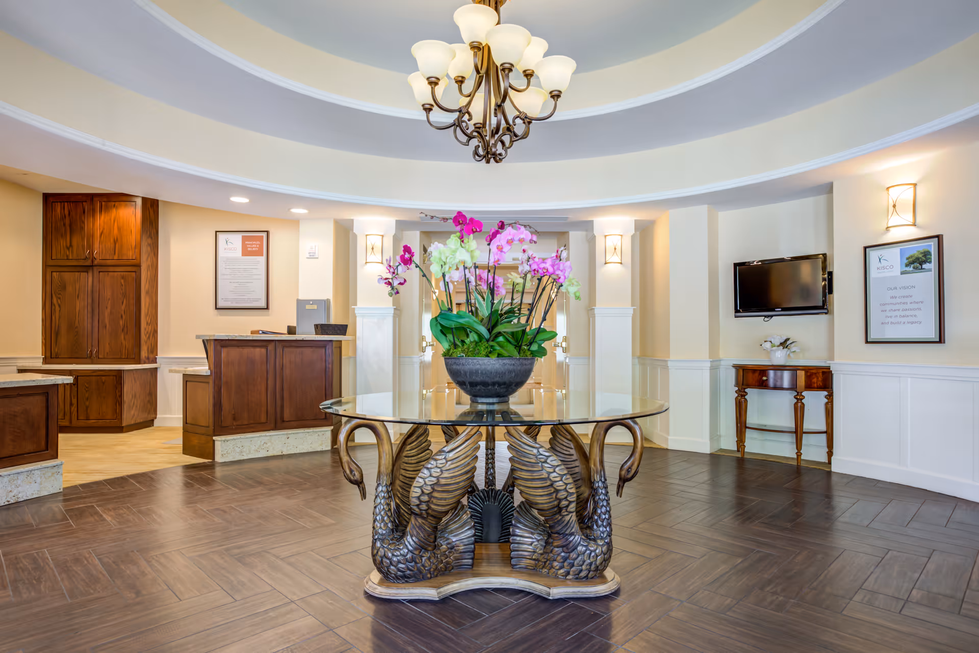 A spacious and elegant lobby area with a round glass table featuring a decorative base shaped like swans. The table holds a large flower arrangement with pink and white orchids. The room has a high ceiling with a chandelier, wood flooring, and cream-colored walls with white wainscoting. There is a reception desk with wooden cabinetry on the left and a wall-mounted TV on the right, along with framed artwork and a small wooden side table.