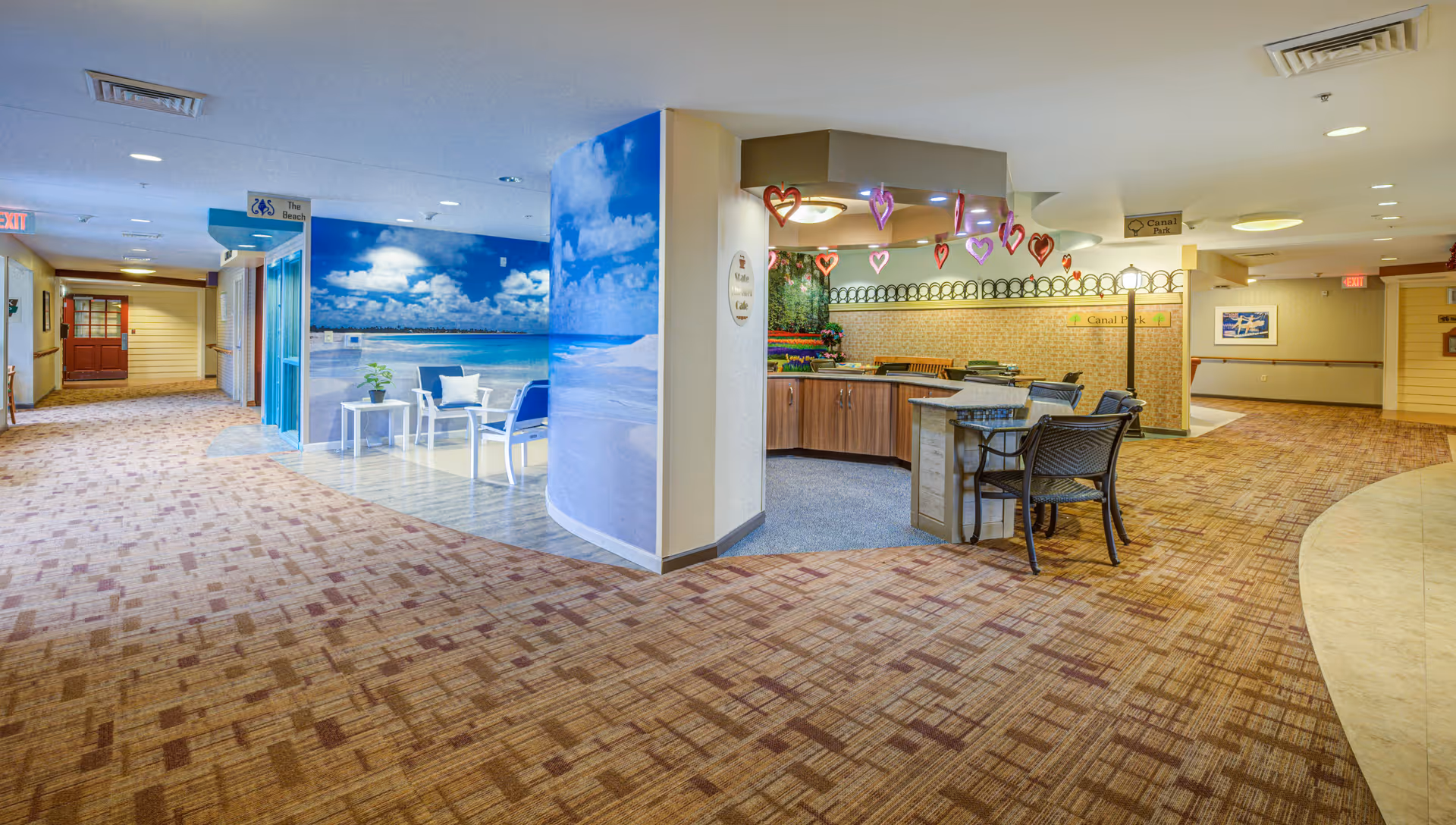 Interior hallway of Arden Courts - ProMedica Memory Care Community with a seating area featuring a beach mural on the wall, a small table with chairs, and a reception or activity desk decorated with hanging heart ornaments. The hallway is carpeted with a patterned design and has directional signs on the ceiling.