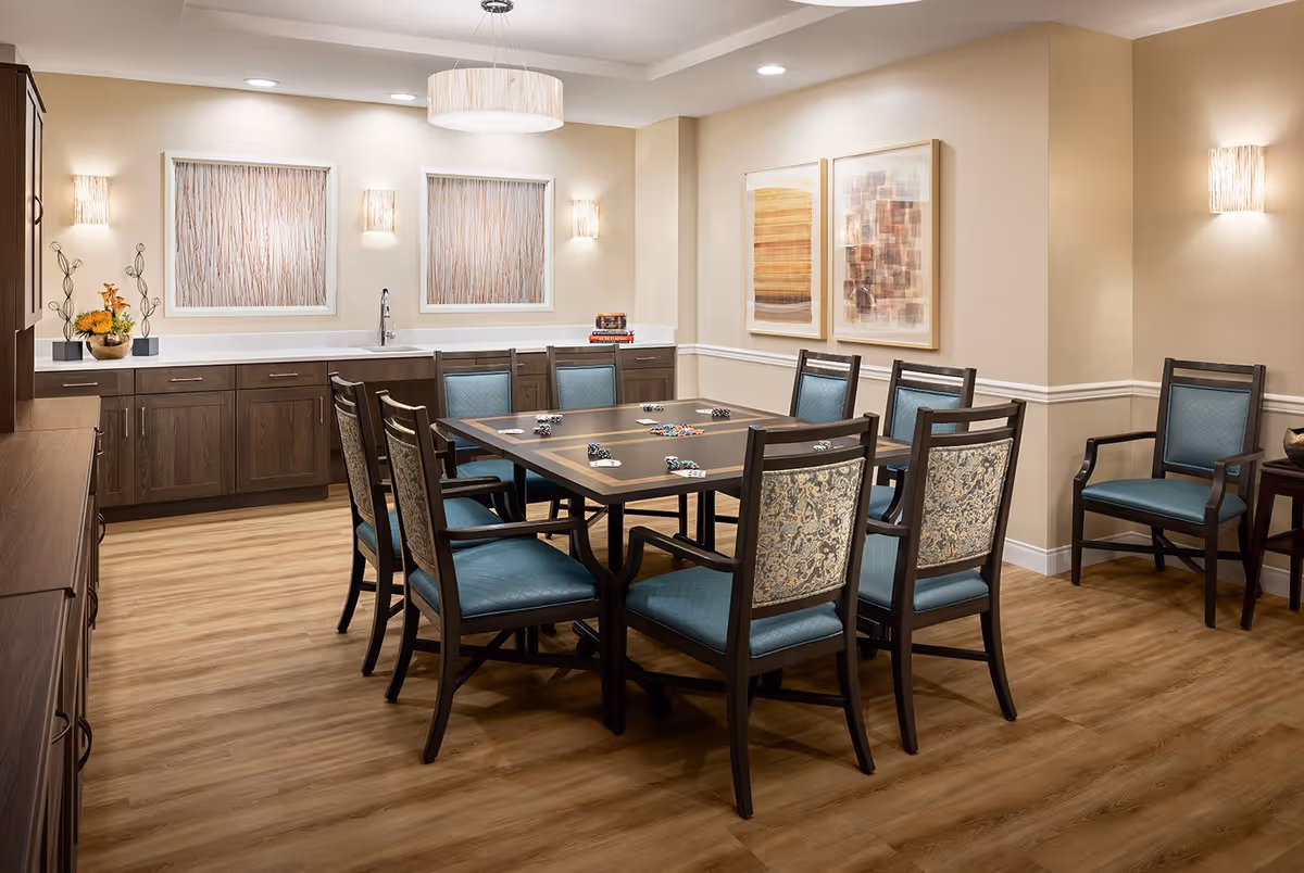 A well-lit room with a square table in the center surrounded by eight wooden chairs with blue cushioned seats and patterned backs. The table has poker chips and playing cards on it. The room features wood flooring, beige walls with white trim, two framed abstract paintings, and two square decorative wall hangings above a countertop with a sink and dark wood cabinets. There are modern wall sconces and a ceiling light fixture providing warm lighting.