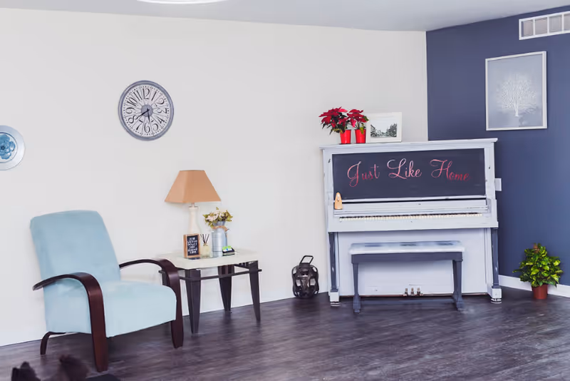 Sitting area with a light-blue armchair, side table and a white upright piano labeled "Just Like Home" against a two-tone wall.