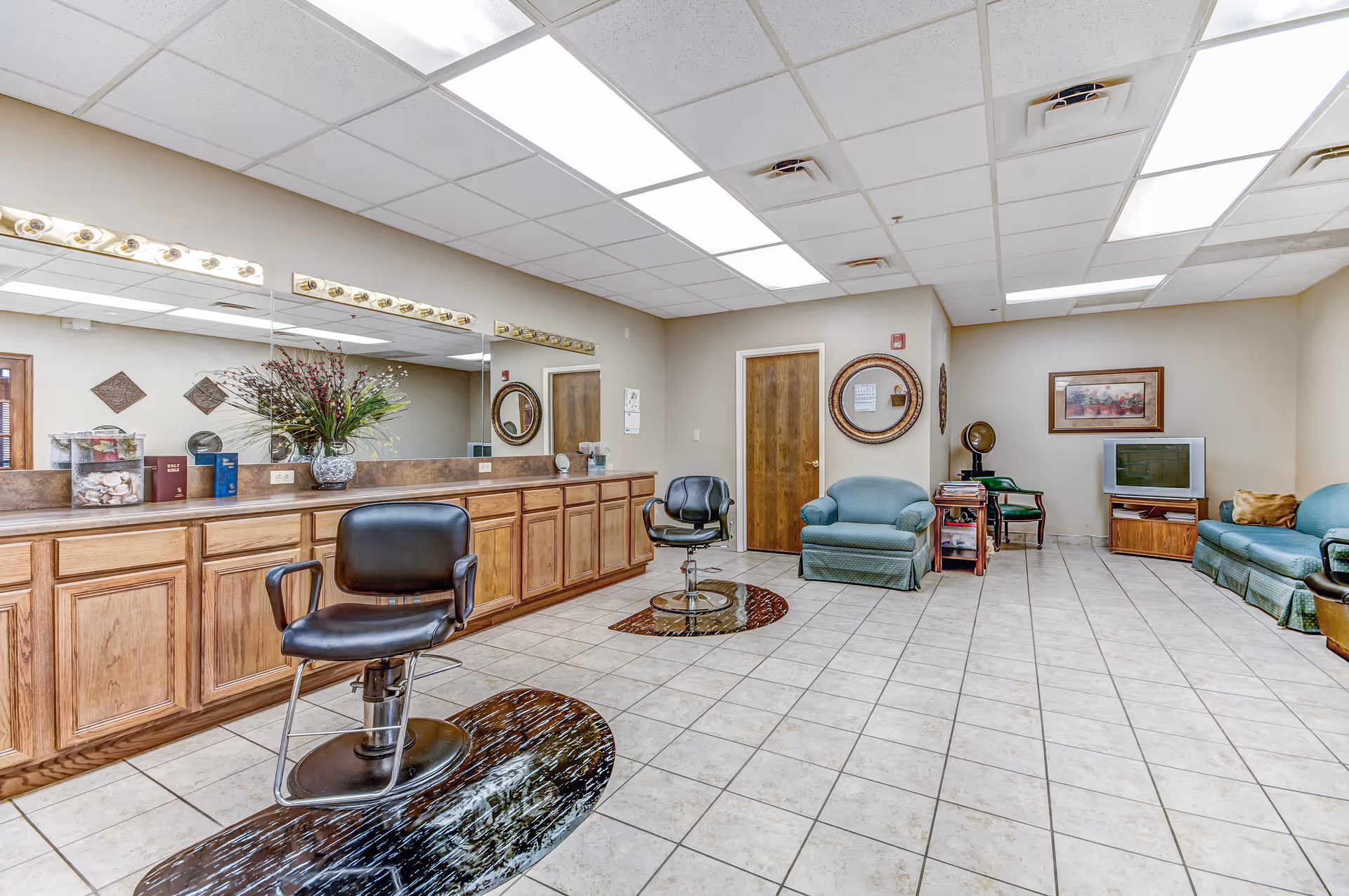 A spacious room with two black salon chairs on circular mats in front of a long counter with wooden cabinets and a large mirror. The room also features a seating area with green upholstered chairs and a couch, a small TV on a wooden stand, and decorative wall hangings. The floor is tiled and the ceiling has fluorescent lighting panels.