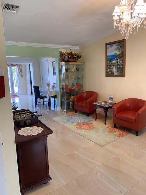 Bright interior seating area with two red armchairs, a floral rug, glass display cabinet and a small table under a chandelier with a dining area visible beyond.