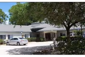 Single-story white senior living building with a covered entrance, a parked car, and trees lining the driveway.