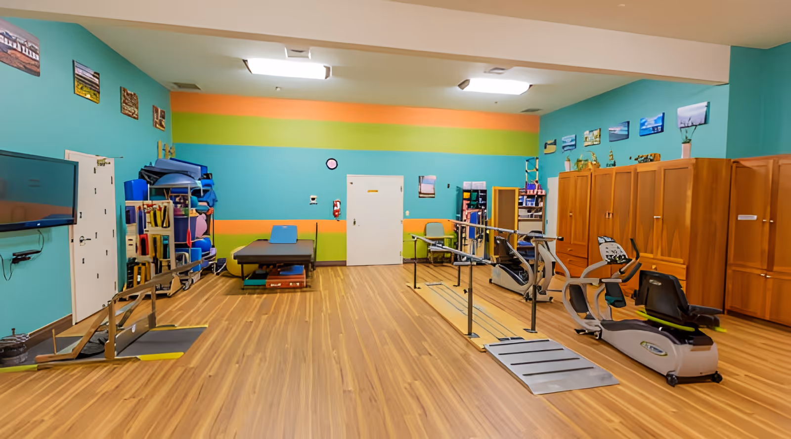 Brightly colored rehabilitation/exercise room with parallel bars, cardio machines, a therapy table, storage lockers, and a wall-mounted TV.