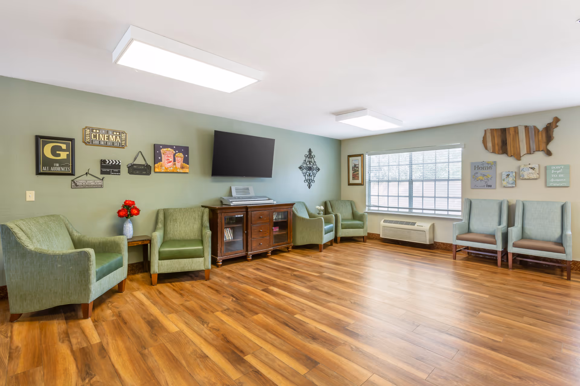 A bright and spacious living room with wooden flooring, light green and blue armchairs arranged along the walls, a wooden cabinet with a flat-screen TV mounted above it, and decorative wall art including a wooden map of the United States and various framed pictures. A large window lets in natural light, and there is an air conditioning unit below the window.