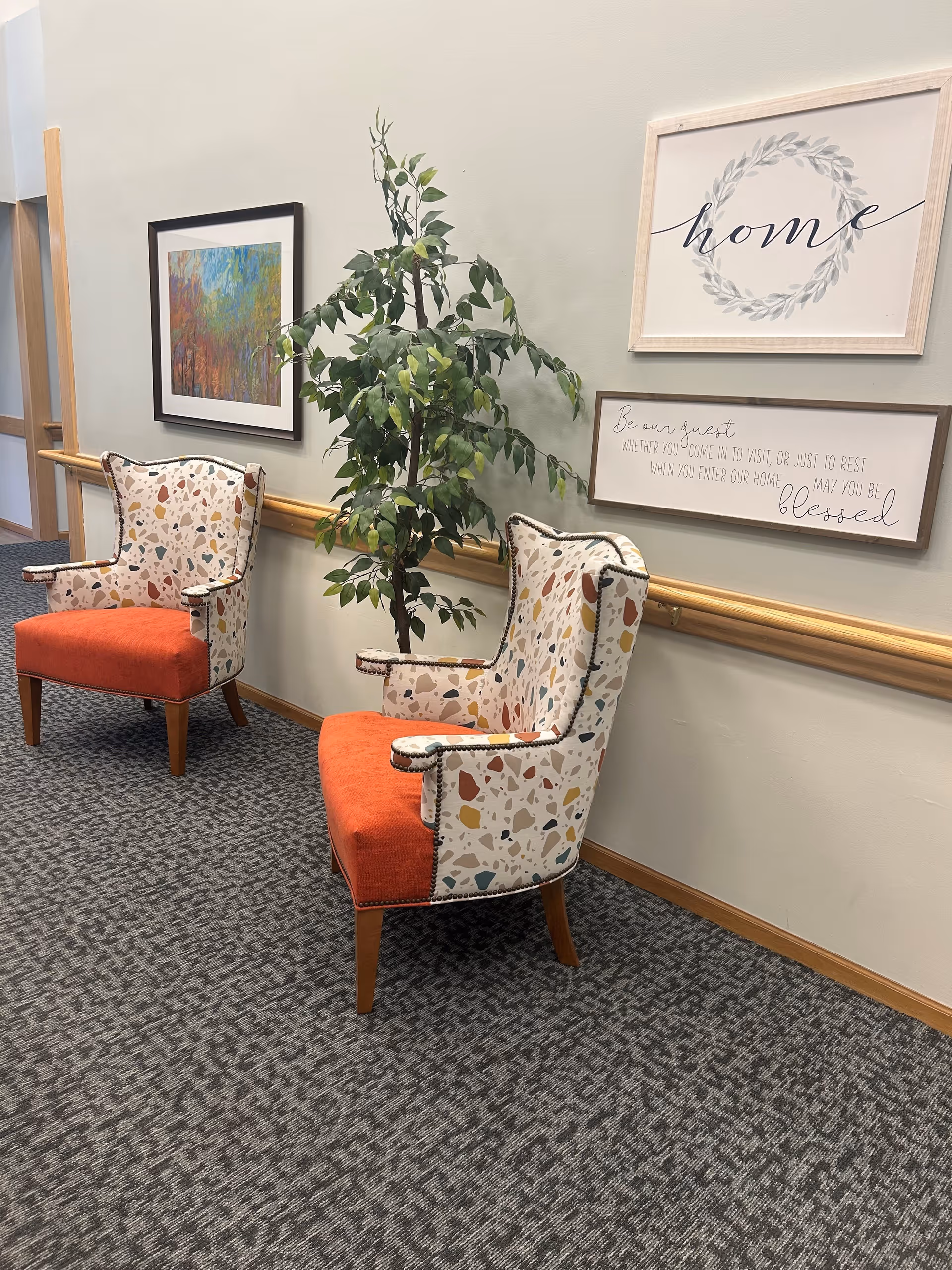 Two patterned armchairs with orange seats are placed against a light-colored wall with a wooden handrail. Between the chairs is a tall green potted plant. On the wall above the chairs are two framed artworks, one with the word 'home' and a wreath design, and the other with a welcoming message. The floor is carpeted with a gray patterned design.