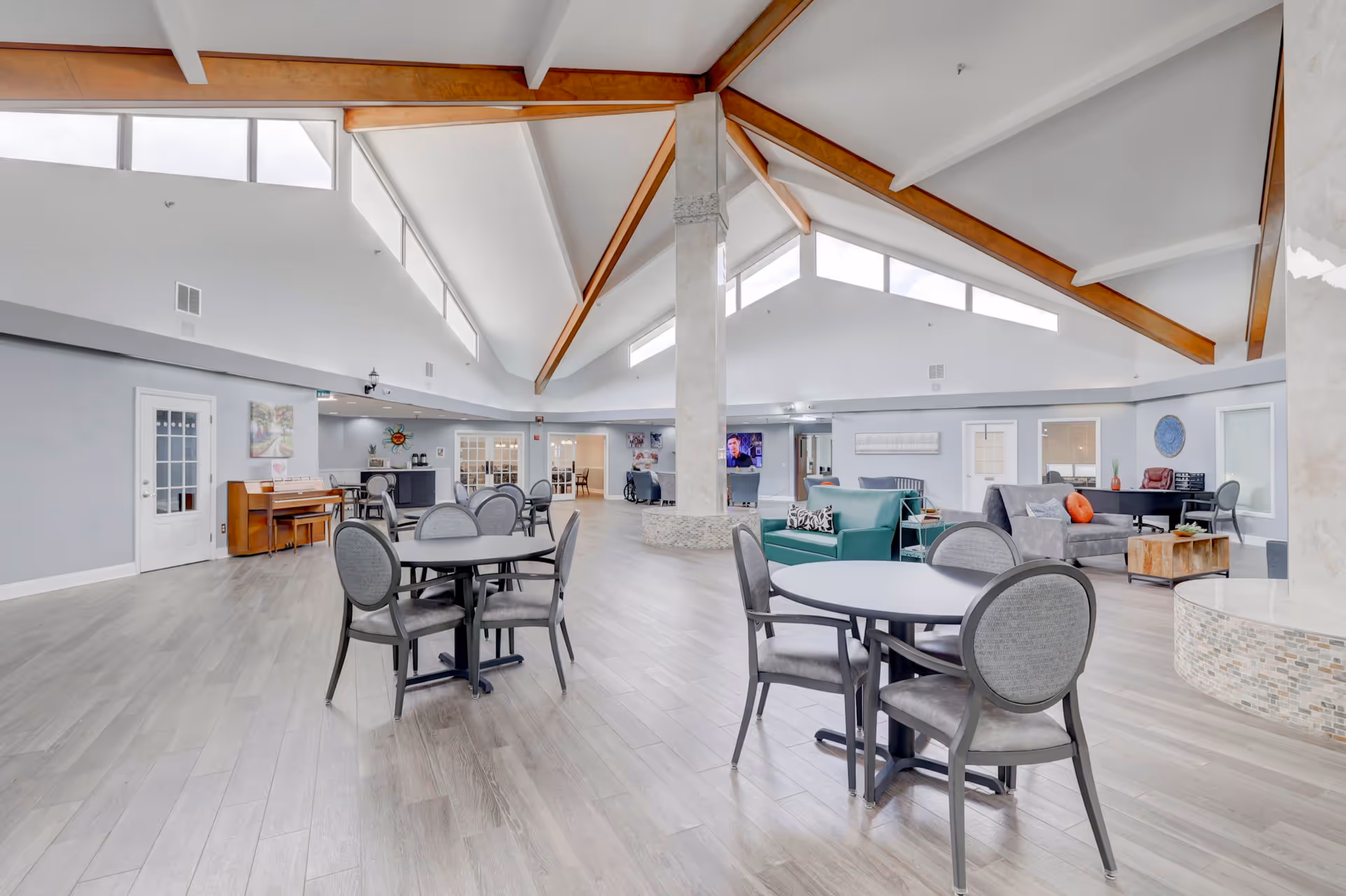 Bright, open senior living common area with round tables and lounge seating under vaulted ceilings with exposed wooden beams.