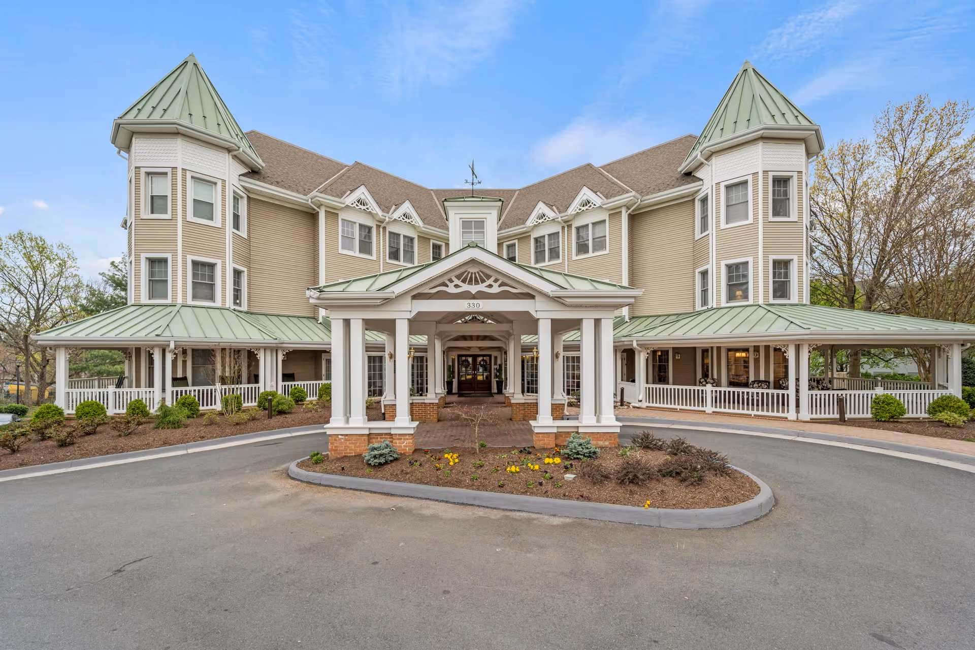 Front exterior of a three-story senior living building with a covered porte-cochère, green metal turrets, wraparound porch, and circular driveway.