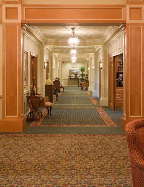 A long, elegant hallway in a senior living facility with patterned carpet, wooden paneling, and multiple light fixtures on the ceiling. The hallway is decorated with chairs, small tables, plants, and framed artwork on the walls.